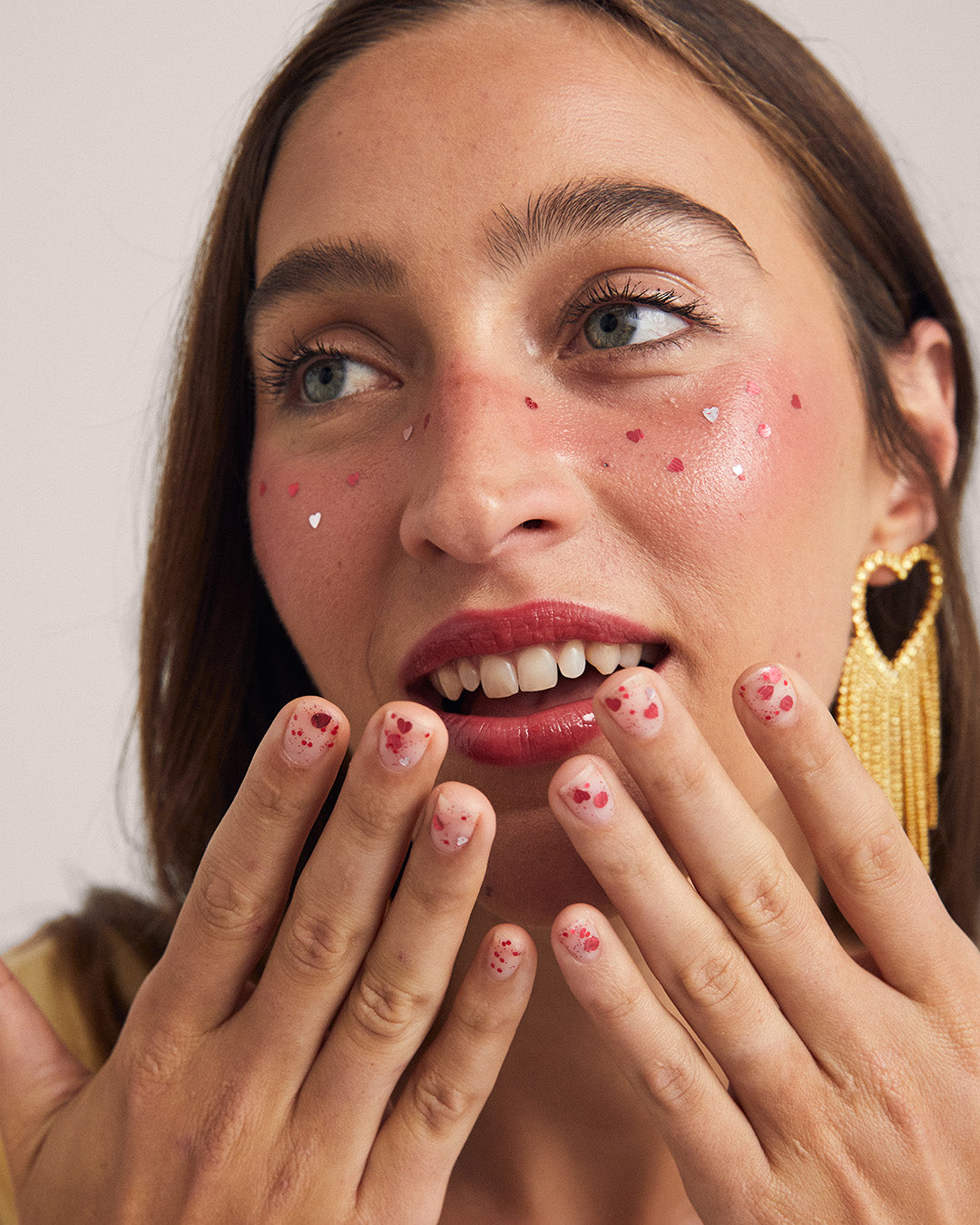A close-up lifestyle shot of a woman with heart-shaped face gems and red heart nail decals, wearing a gold heart earring against a neutral light gray background.