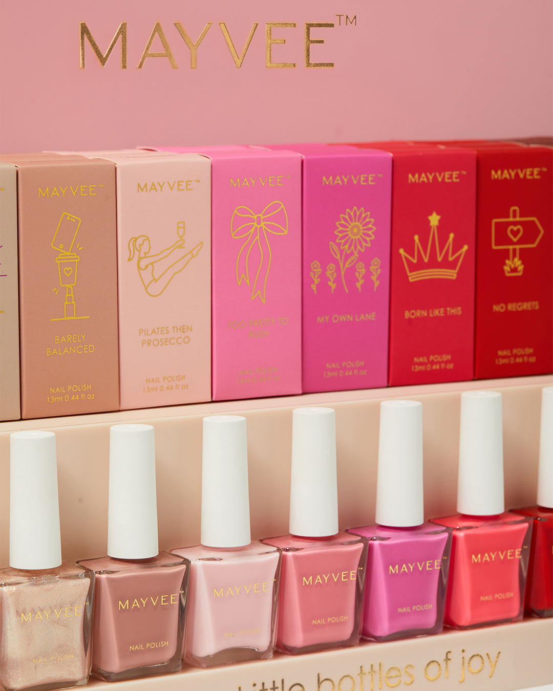 Mayvee nail polish set with multiple pink and red boxes and matching bottles displayed on a pink shelf. The boxes feature gold line-art icons and the brand name MAYVEE is printed on the top and bottles.